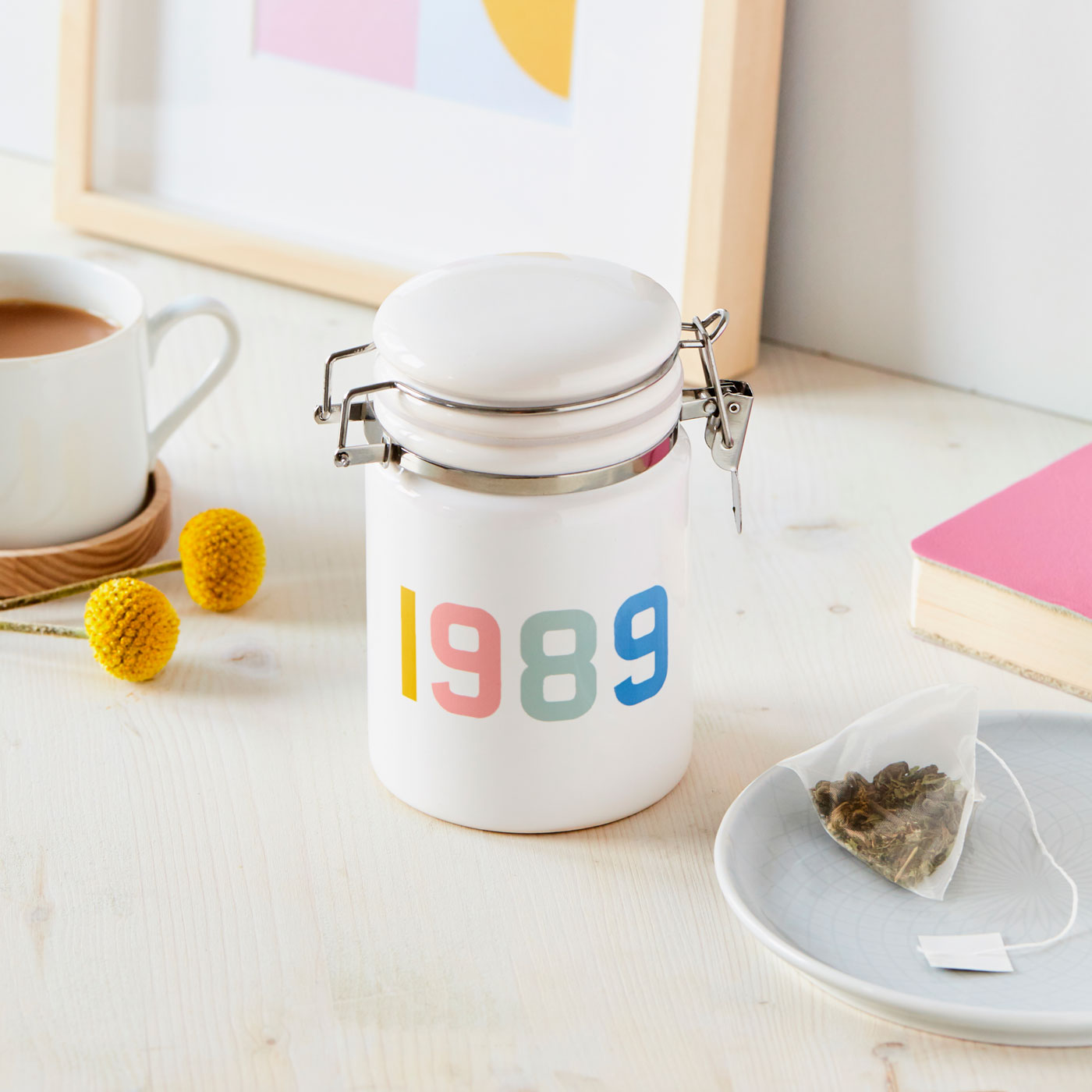 White jar with '1989' on a table with a cup of coffee and tea bag.