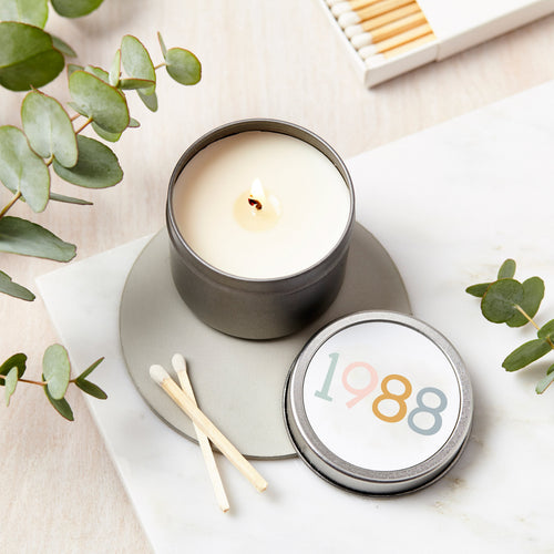 Candle in a tin with '1988' label on a light surface with eucalyptus leaves.