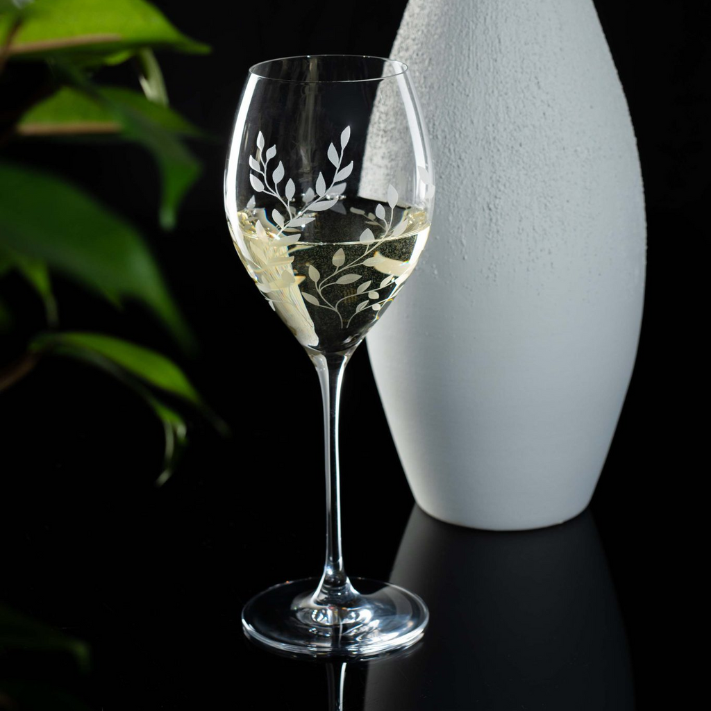Etched wine glass with white wine next to a white vase on a dark surface