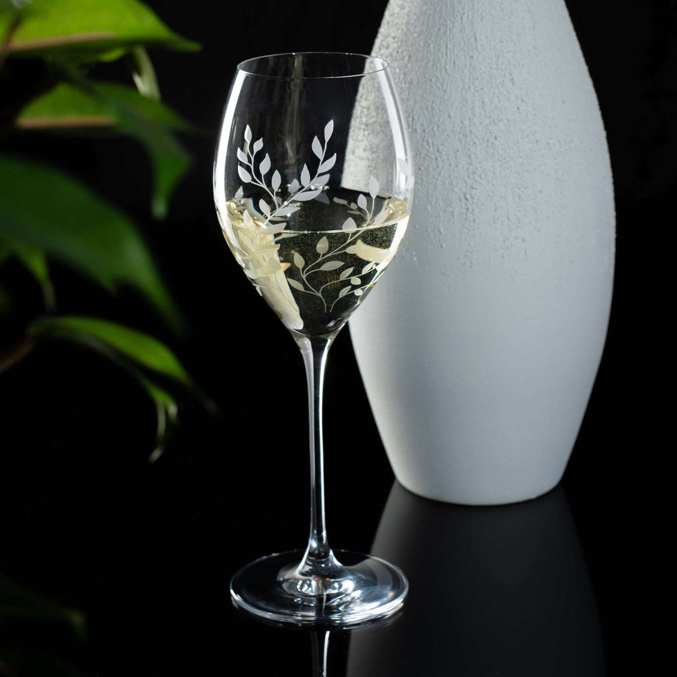 Etched wine glass with white wine next to a white vase on a dark surface