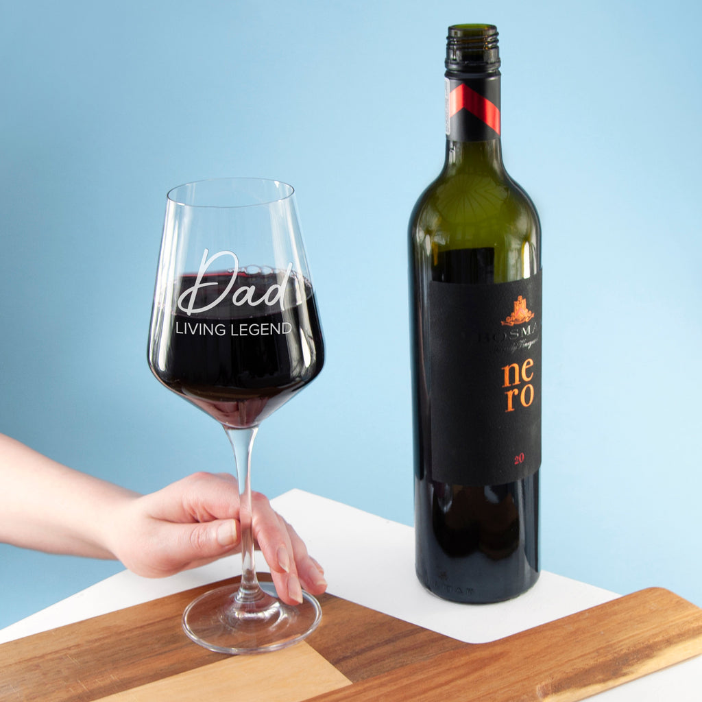 Wine glass with 'Dad Living Legend' text and wine bottle on a wooden board against a blue background