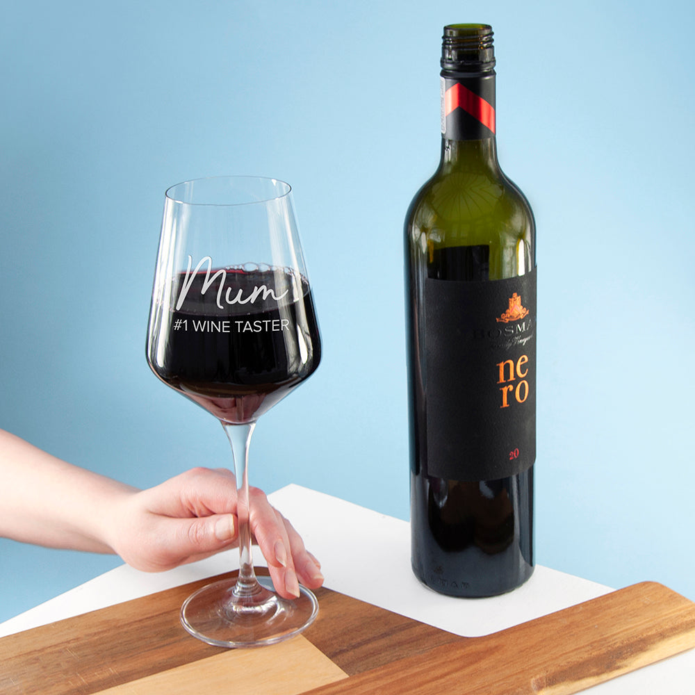 Wine glass with 'Mum #1 Wine Taster' text and a bottle of wine on a wooden board against a blue background