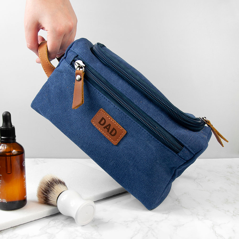 Blue toiletry bag with 'DAD' patch held by a hand, next to a bottle and brush on a marble surface.