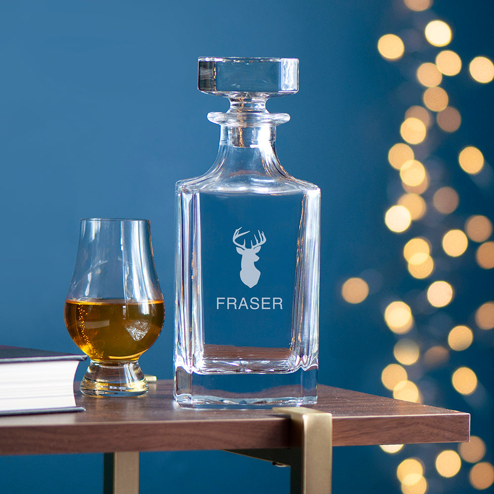 Clear glass decanter with 'Fraser' branding next to a glass of whiskey on a wooden surface with a blurred festive background.