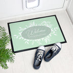 Personalized doormat with floral design and text, placed on a light wooden floor with shoes and a plant.