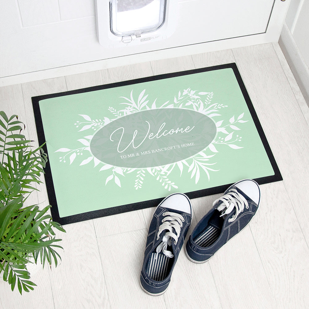 Personalized doormat with floral design and text, placed on a light wooden floor with shoes and a plant.