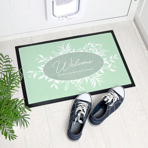 Personalized doormat with floral design and text, placed on a light wooden floor with shoes and a plant.