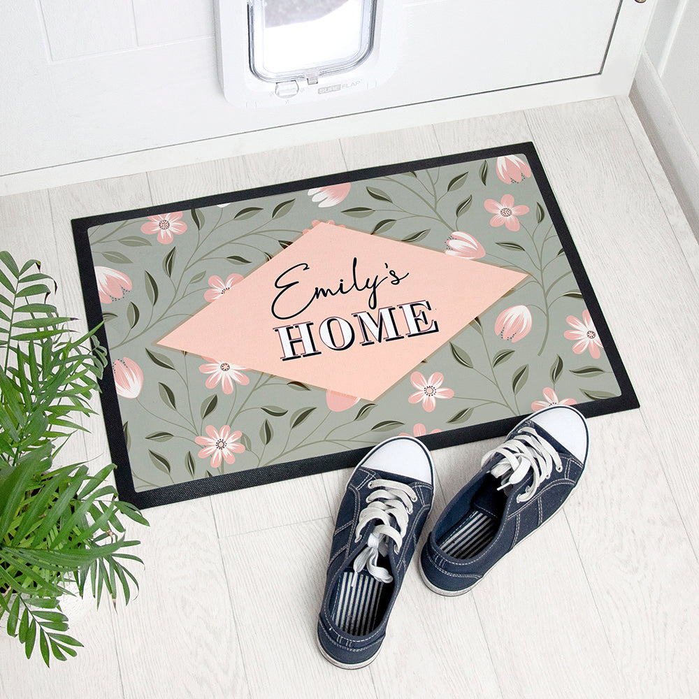 Personalized doormat with floral design and 'Emily's Home' text, placed on a wooden floor with shoes and a plant.