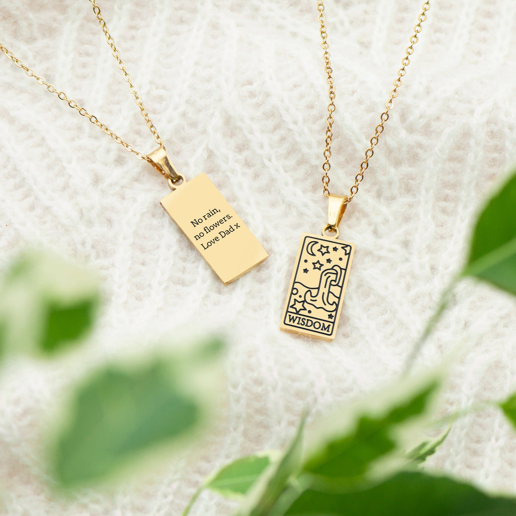 Two gold necklaces with pendants on a textured white surface with green leaves in the background.