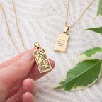 Gold pendant with 'WISDOM' text held by a hand, with another pendant and green leaves in the background.