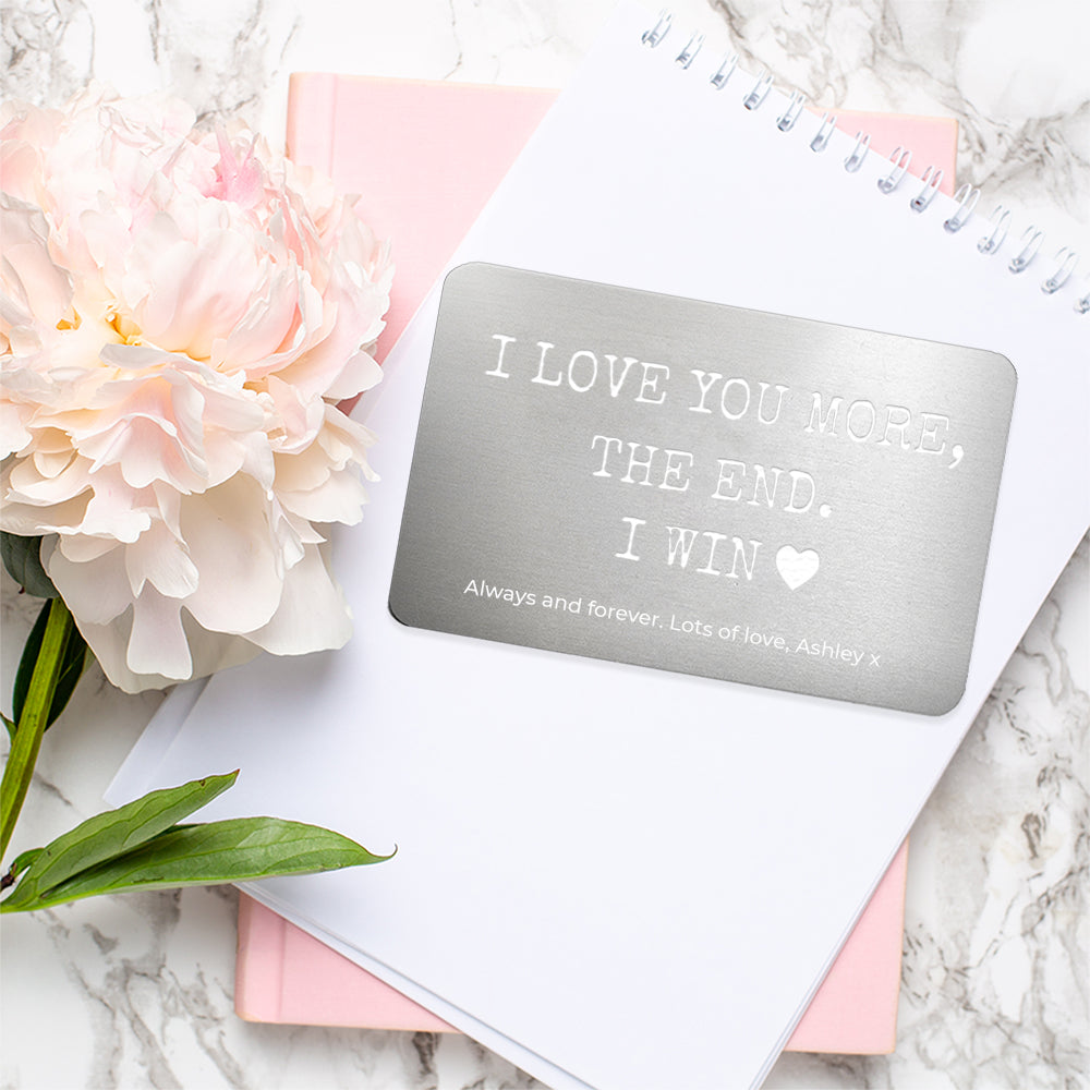 Card with 'I LOVE YOU MORE, THE END. I WIN' text on a marble surface with flowers