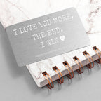 Notebook with a card inside reading 'I love you more, the end. I win.' on a marble surface.