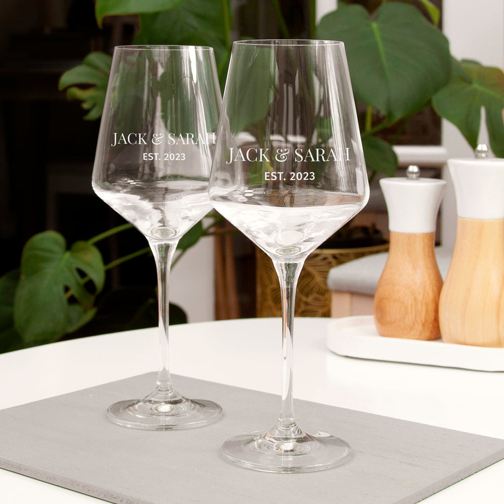 Two personalized wine glasses on a table with plants in the background
