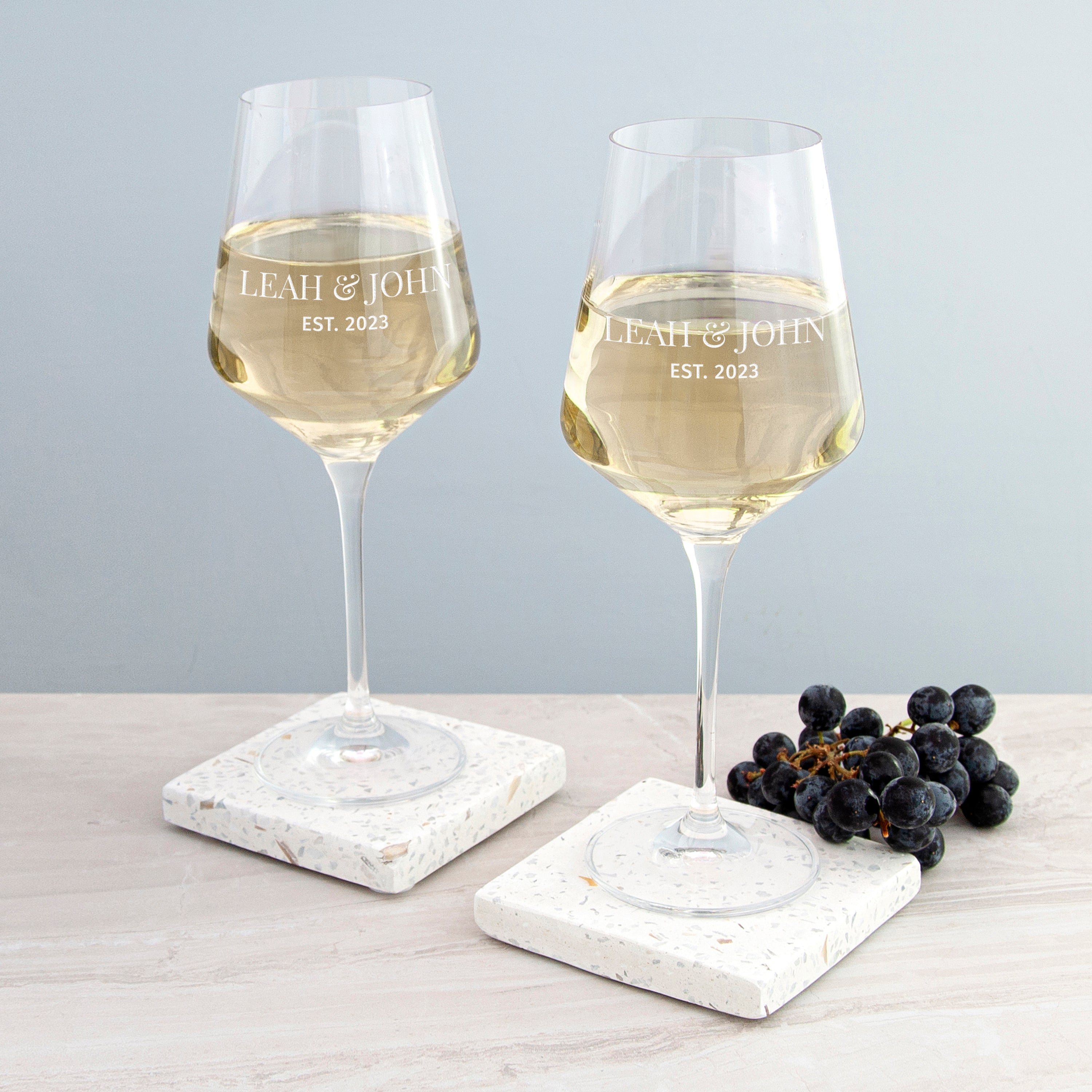 Two wine glasses with white wine on marble coasters, with 'LEAH & JOH' and 'EST. 2023' text.