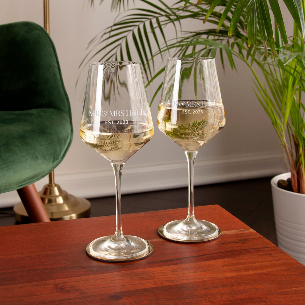 Two wine glasses with engraved text on a wooden table, surrounded by a plant and green chair.
