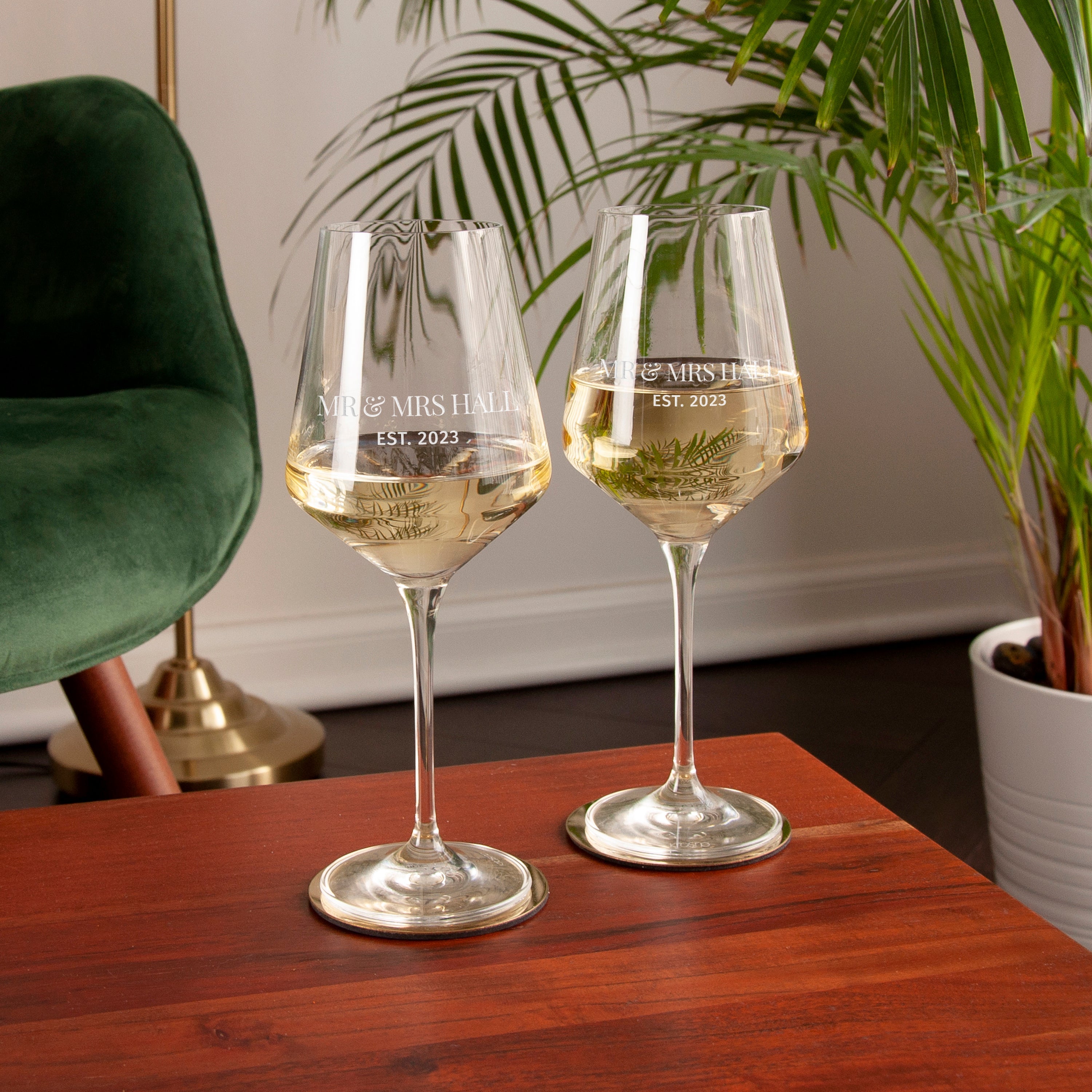 Two wine glasses with engraved text on a wooden table, surrounded by a plant and green chair.
