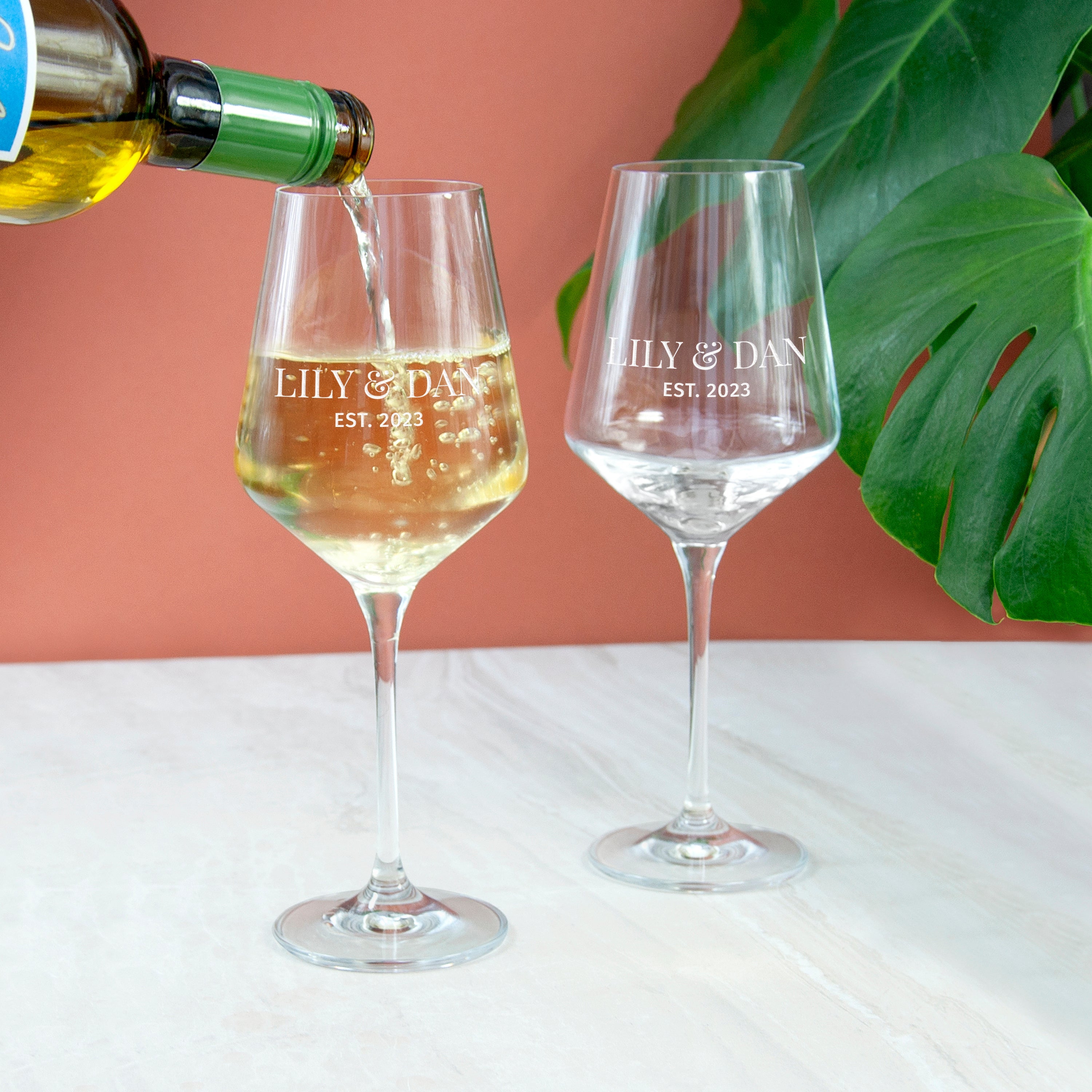 Wine glasses with 'ILY & DAN EST. 2023' engraving being filled with white wine against a pink background.