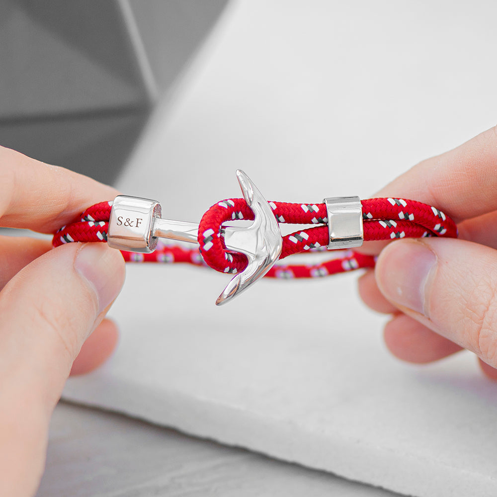 Red braided bracelet with silver anchor charm held by hands on a light gray background