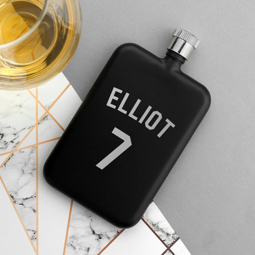 Black flask with 'ELLiot 7' engraving on a marble surface with a glass of whiskey.