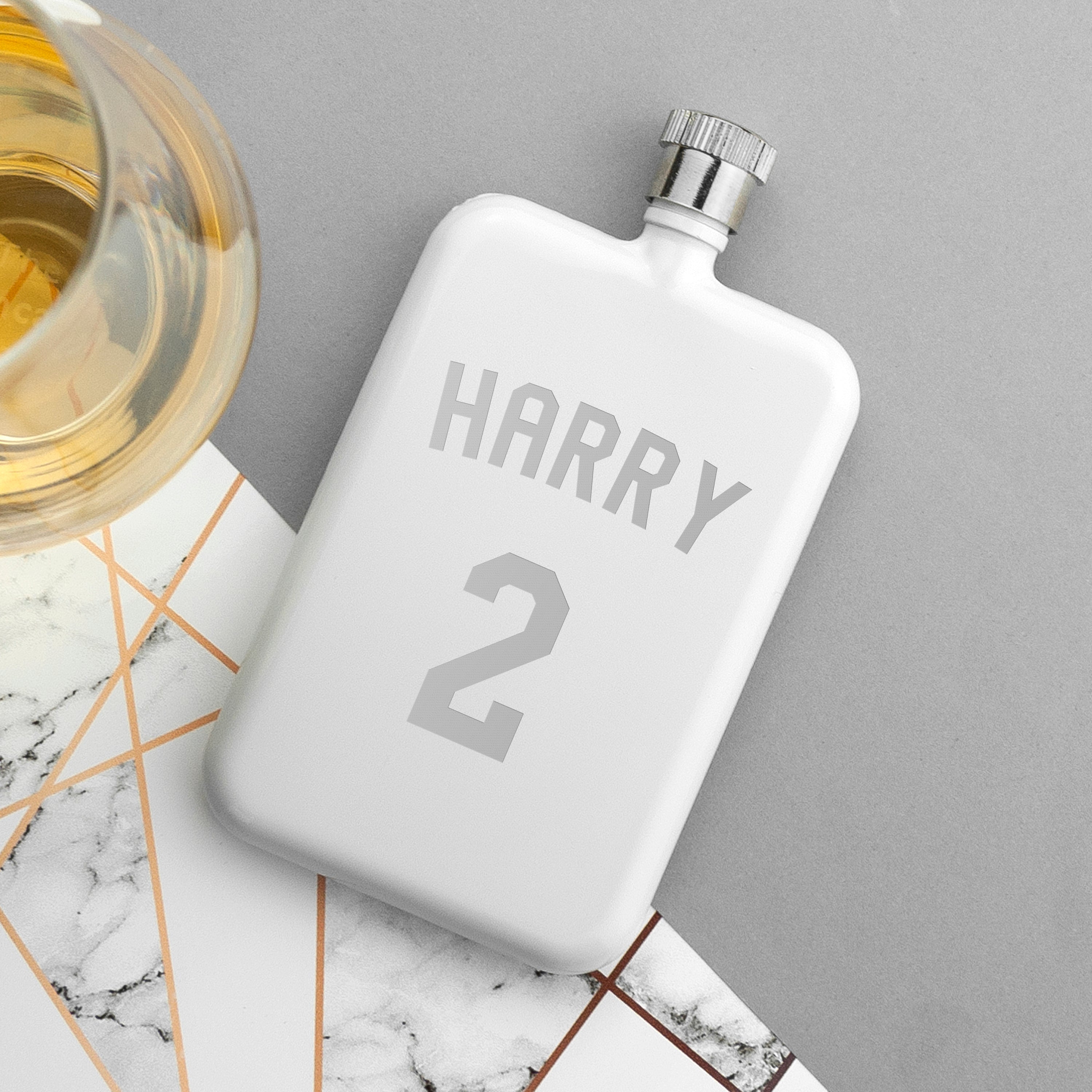 White flask with 'HARRY 2' engraving on a marble surface with a glass of liquid.