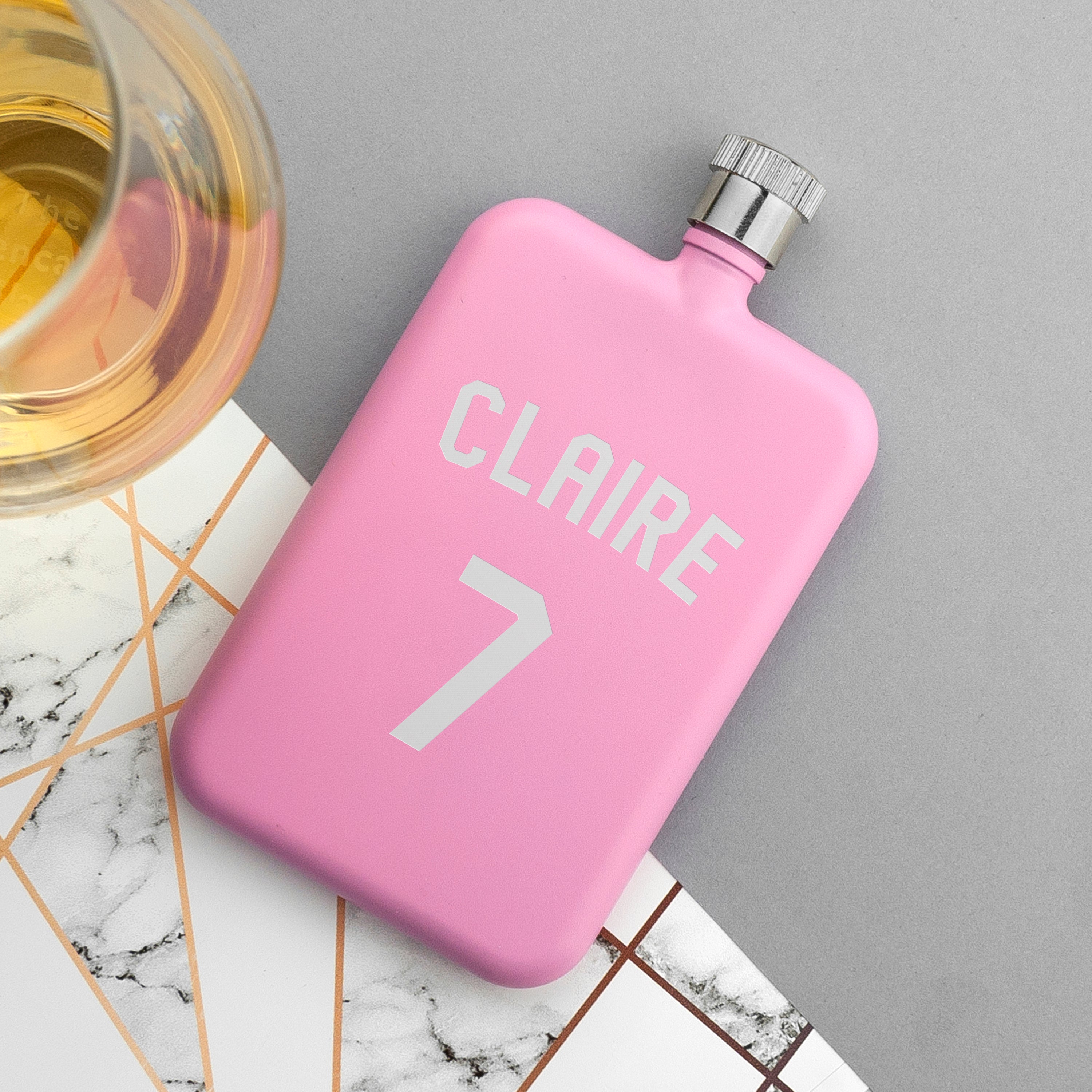 Pink flask with 'CLAIRE' and number '7' on a marble surface with a glass of liquid.