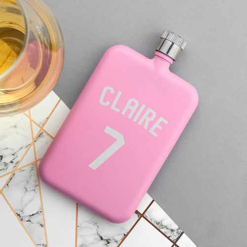 Pink flask with 'CLAIRE' and number '7' on a marble surface with a glass of liquid.