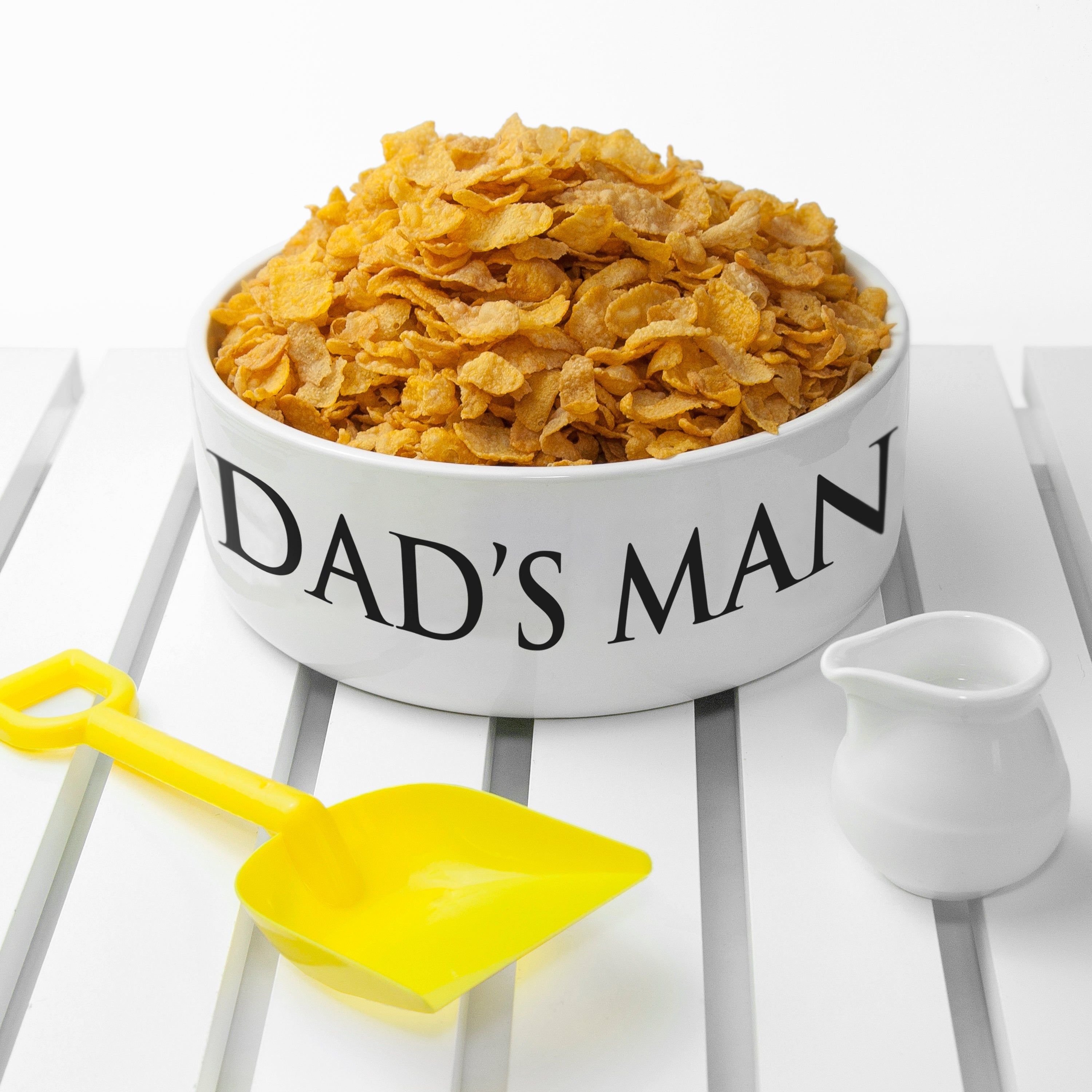 Bowl of cereal with 'DAD'S MAN' text, yellow spoon, and white pitcher on a white background