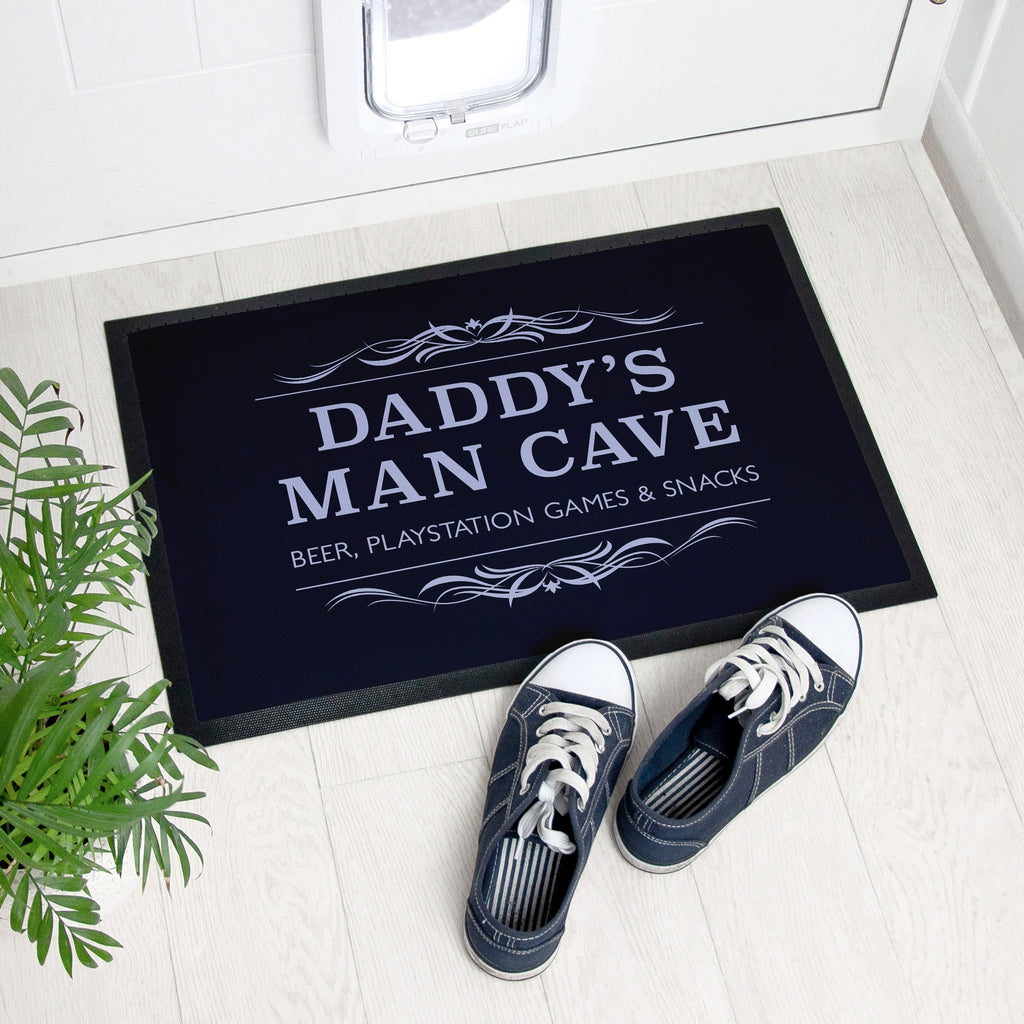 Doormat with 'Daddy's Man Cave' text on a white floor with shoes and plant.