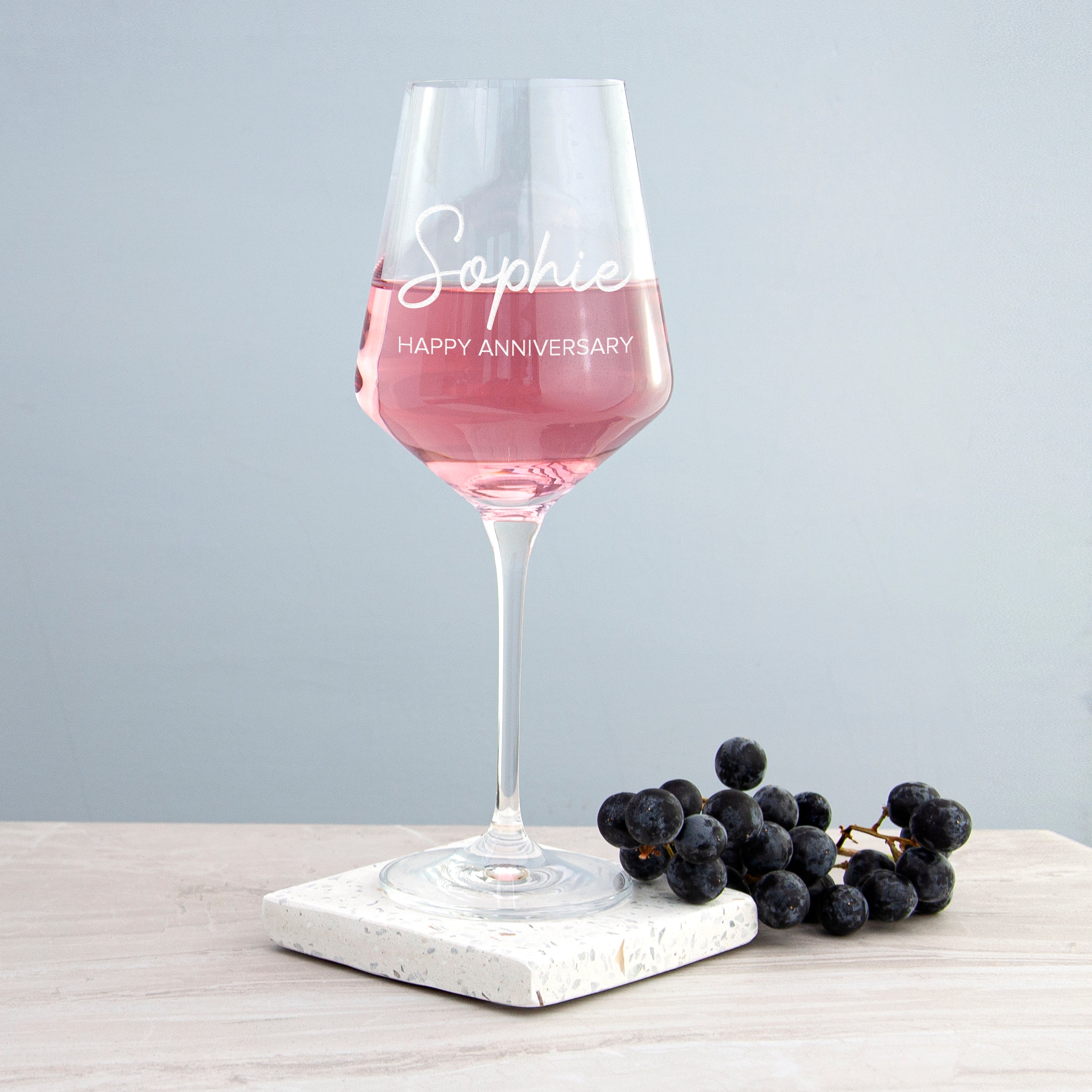 Wine glass with pink liquid on a marble coaster with grapes, against a light gray background