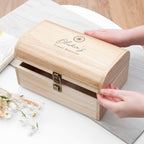 Wooden box with 'Olivia's Travel Memories' engraving on a light surface with hands opening it.