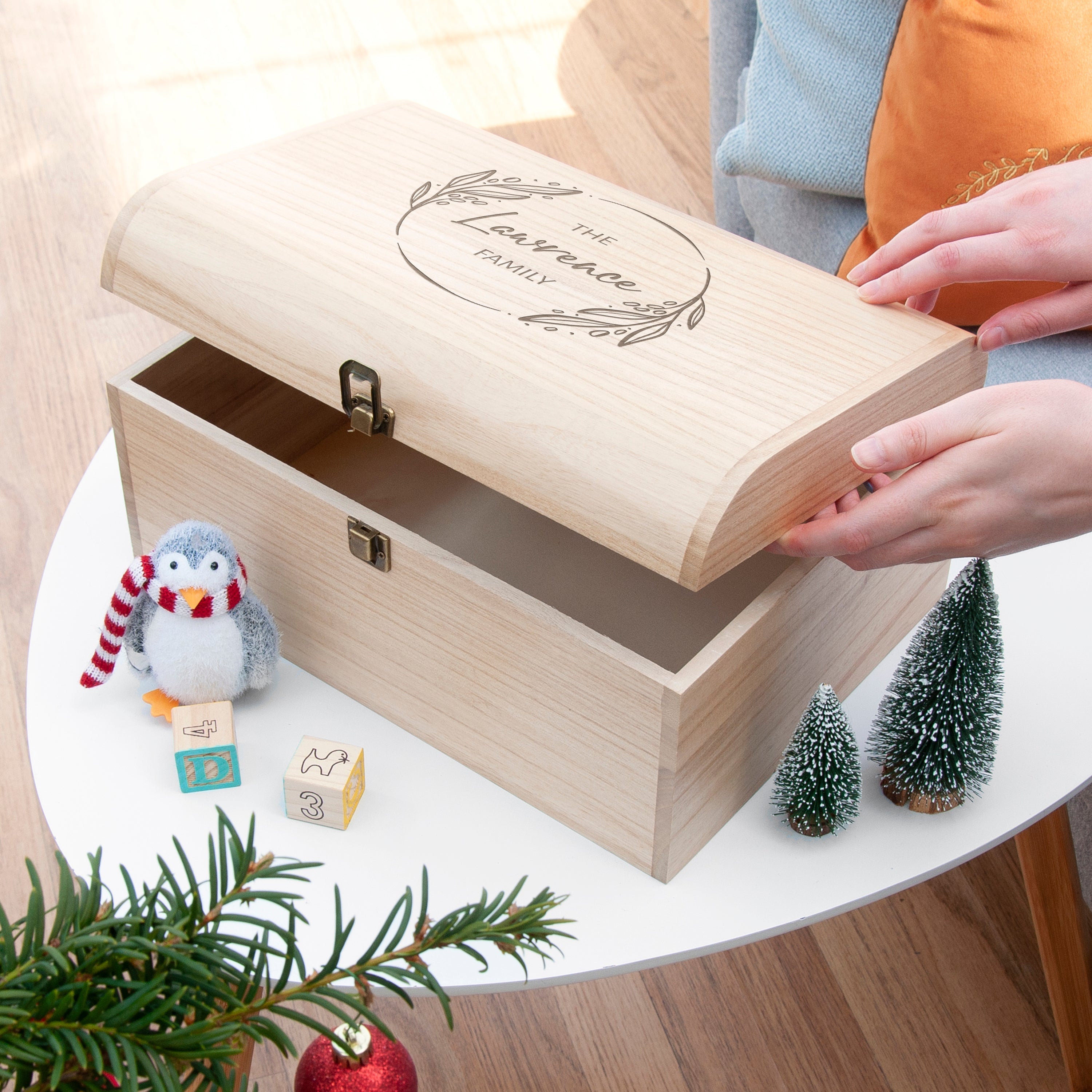 Wooden box with 'Isabelle's First Christmas' engraving, penguin toy, and small Christmas trees on a table.