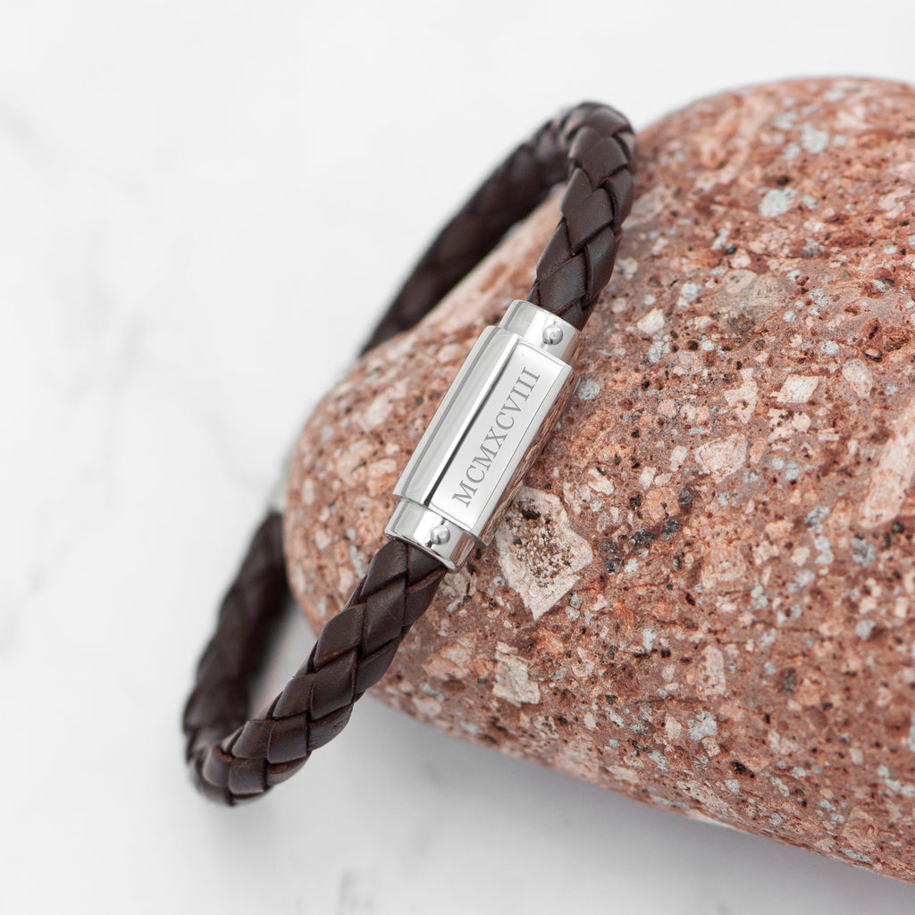 Brown braided leather bracelet with a silver tag on a textured stone surface