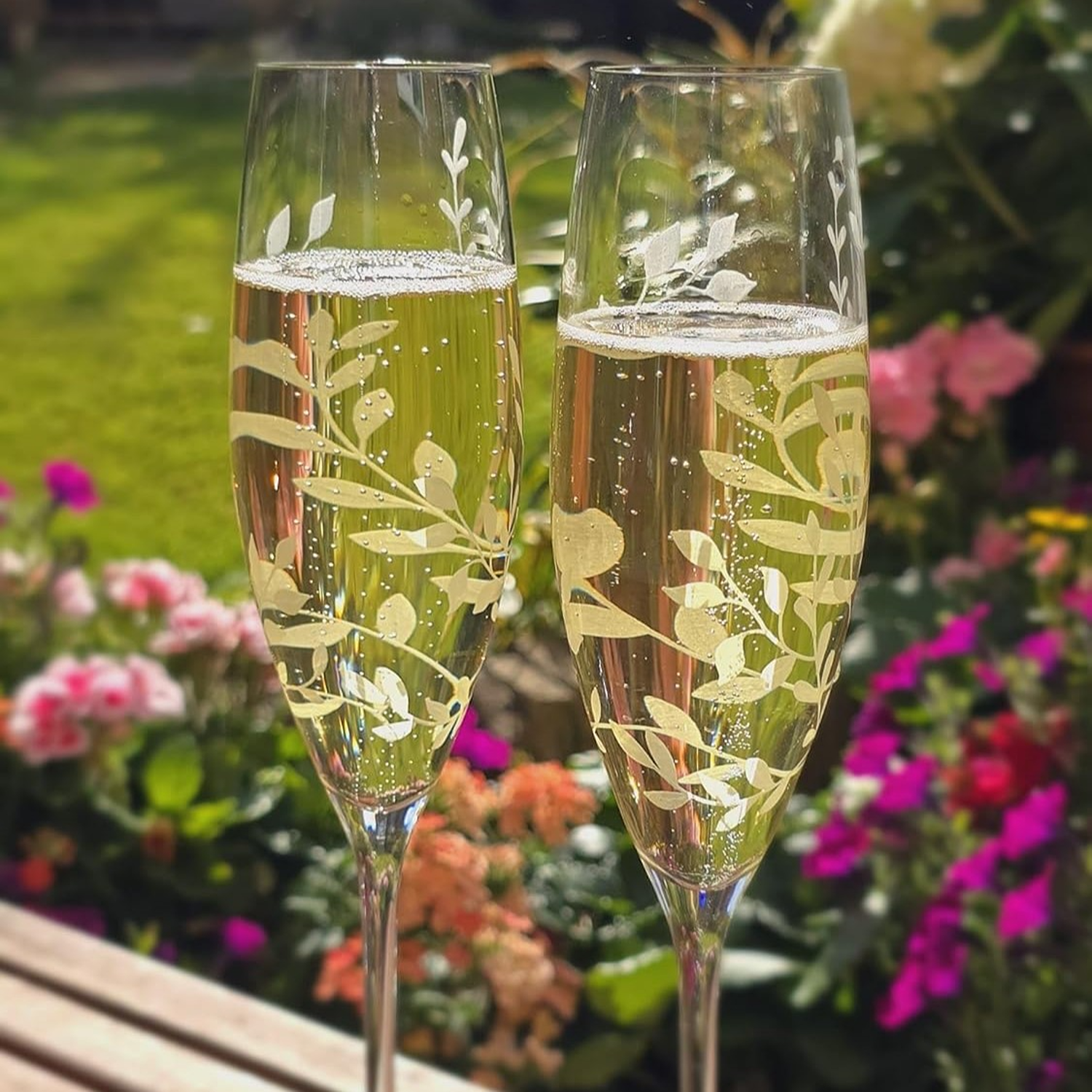 Two etched champagne flutes with a floral design filled with champagne against a garden background.