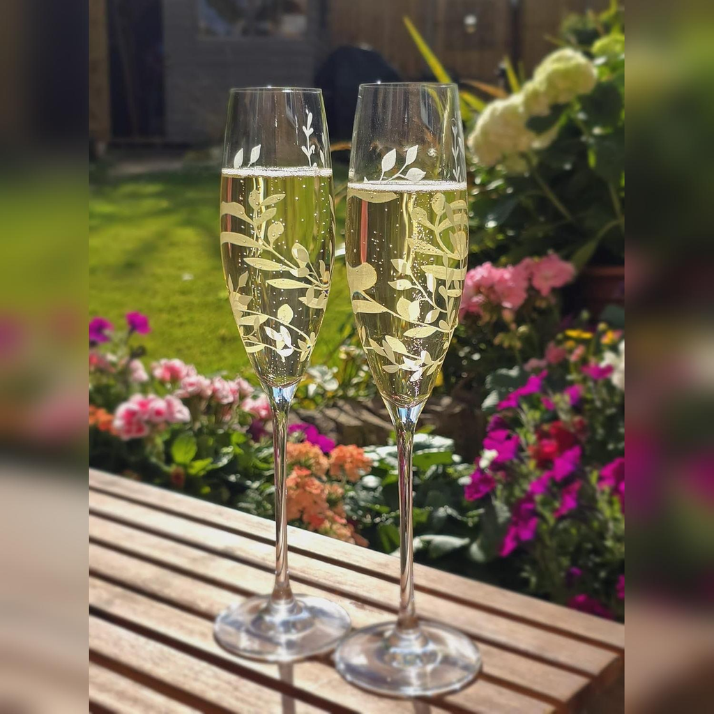 Two etched champagne flutes on a wooden surface with a garden background