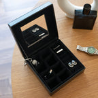 Black jewelry box with mirror lid containing jewelry on a wooden surface.
