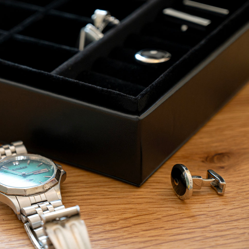 Silver watch and cufflinks on a wooden surface with a black jewelry box in the background.