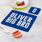 Blue placemat with 'OLIVER BIG BRO' text on a marble surface with fruit and a bowl.
