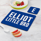 Blue placemat with 'ELLiot LITTLE BRO' text on a marble surface with fruit and a bowl.