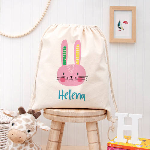 Drawstring bag with a rabbit design and 'Helena' text on a wooden stool, with a giraffe plush toy and books in the background.