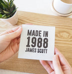 Hand holding a card with 'Made in 1988 James Scott' text on a wooden surface.