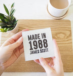 Person holding a card with 'Made in 1988 James Scott' text on a wooden surface with a cup and plant in the background.