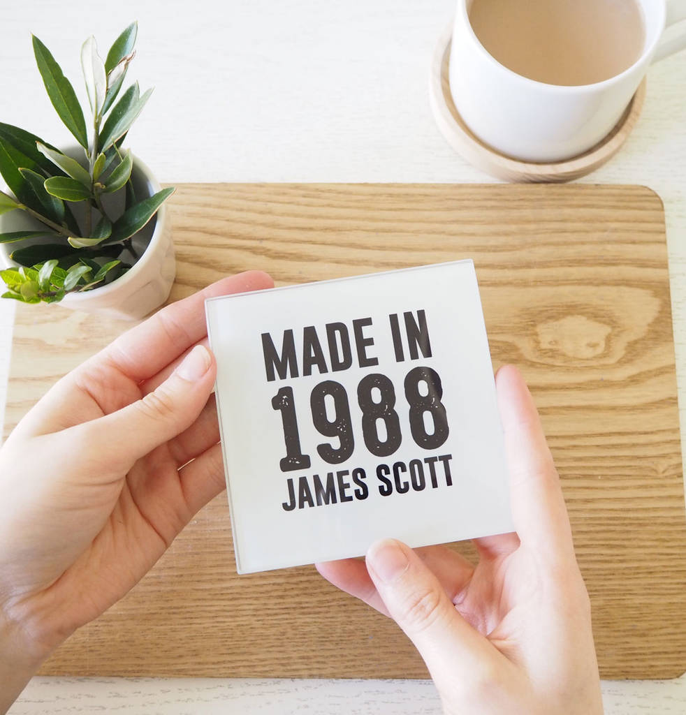 Person holding a card with 'Made in 1988 James Scott' text on a wooden surface with a cup and plant in the background.