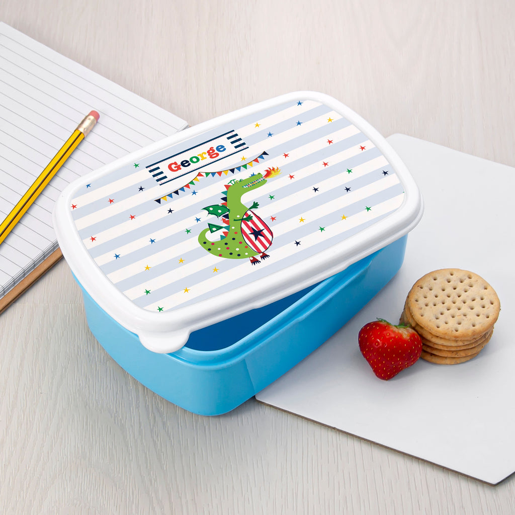 Blue lunch box with a cartoon crocodile design on a white surface with snacks.