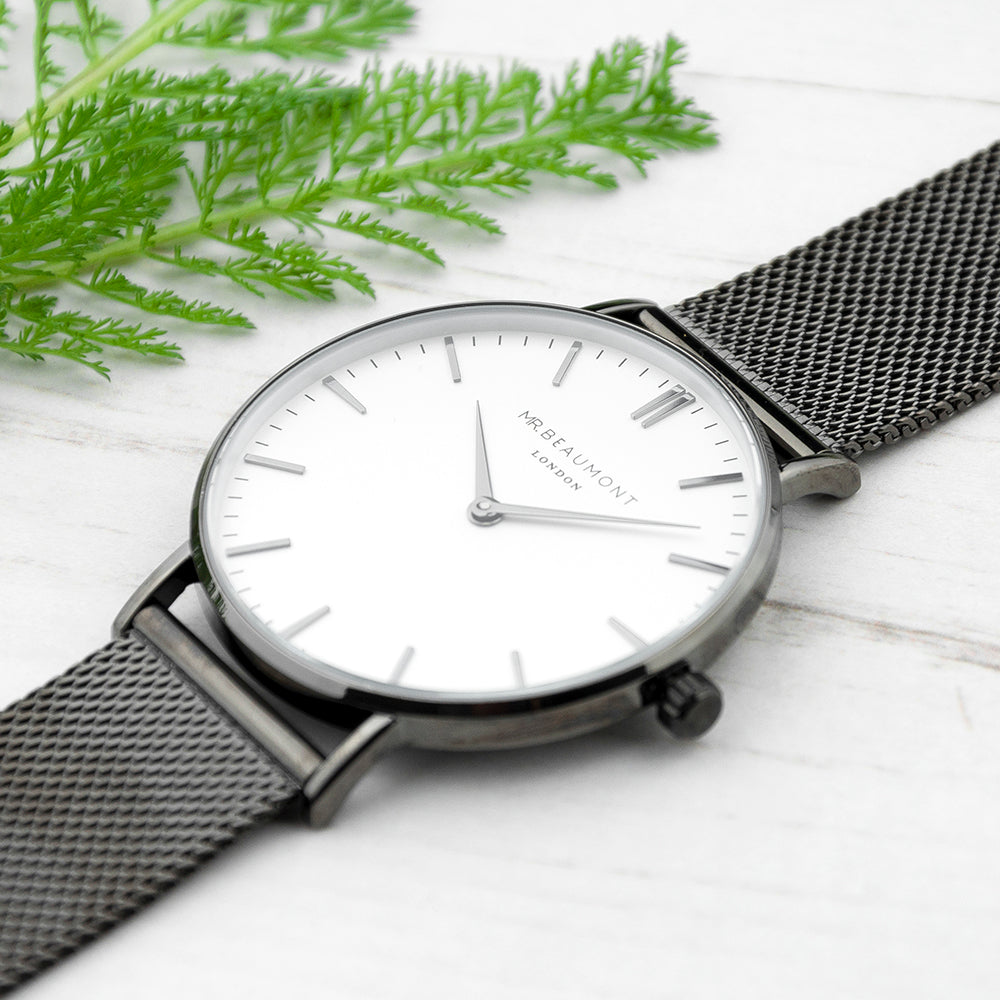 Silver watch with black mesh strap on a white surface with green leaves.
