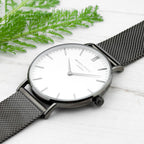 Silver watch with black mesh strap on a white surface with green leaves.