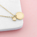 Gold necklace with a heart-shaped pendant and engraved disc on a white and pink background