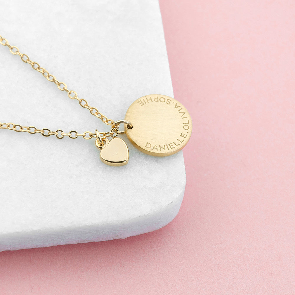 Gold necklace with a heart-shaped pendant and engraved disc on a white and pink background