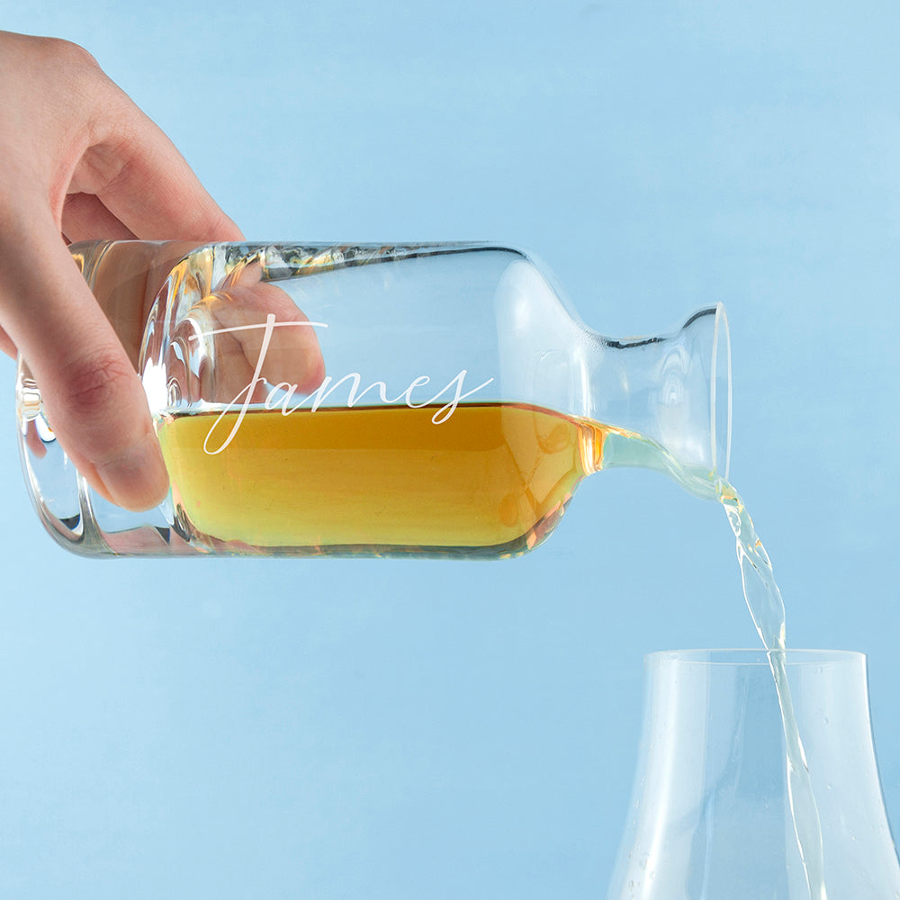 Clear glass bottle with 'James' engraving being poured into a glass against a light blue background