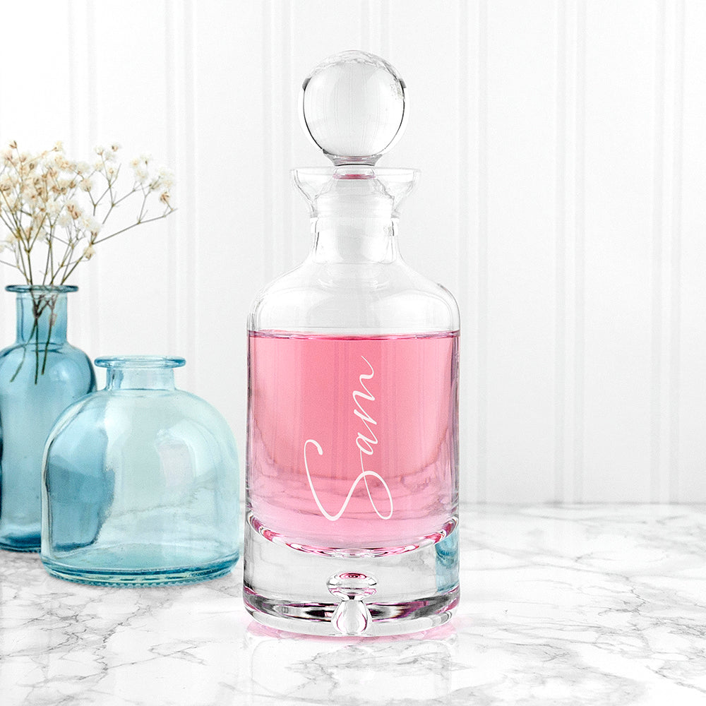 Clear glass decanter with pink liquid and 'Sam' engraving on a marble surface with light blue vases in the background.