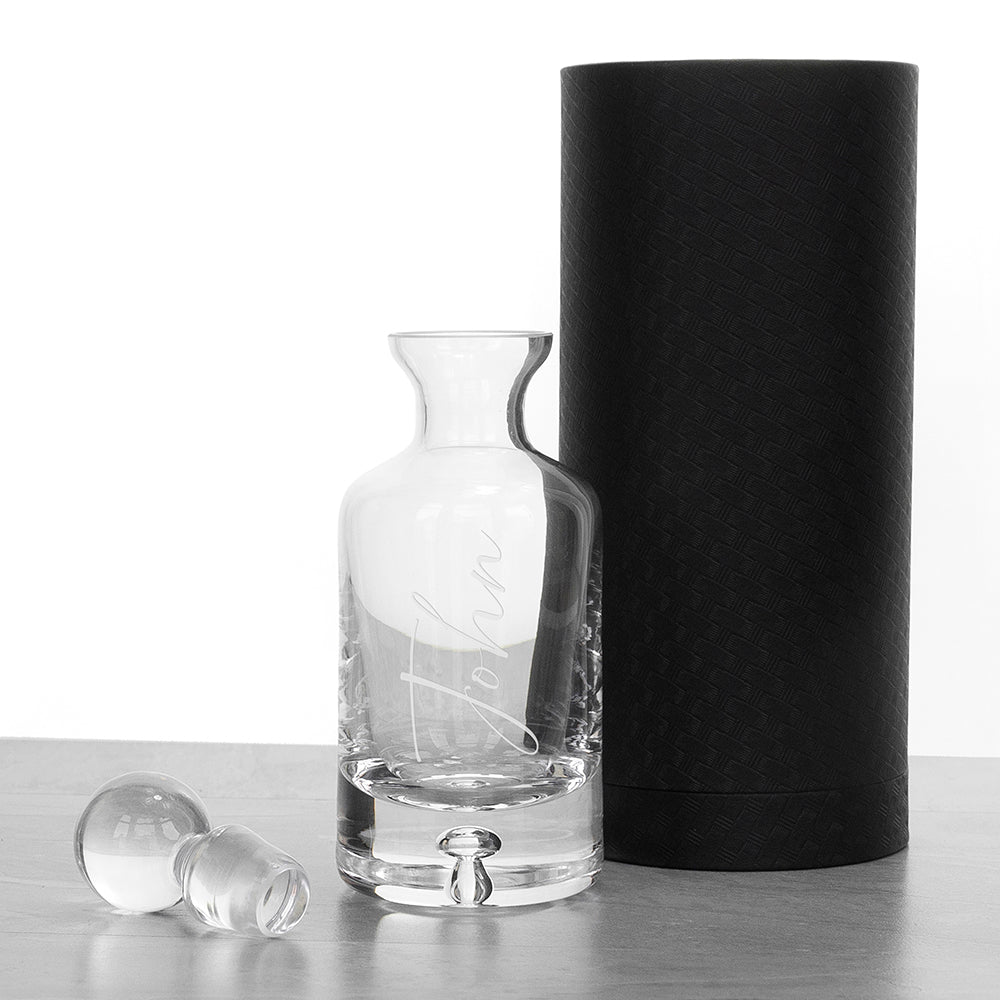 Clear glass carafe with handle and stopper next to a black cylindrical container on a white background